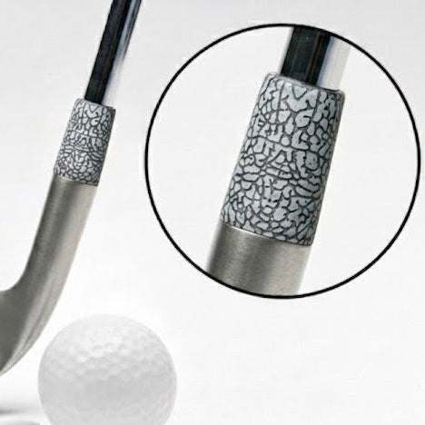 A silver-colored golf club head with a textured black and silver ferrule, shown next to a white golf ball and teeing stick, with a circular inset detail of the grip pattern.