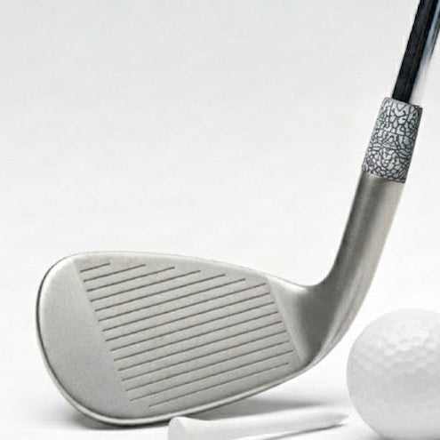 A silver-colored golf club head with a textured black and silver ferrule, shown next to a white golf ball and teeing stick, with a circular inset detail of the grip pattern.