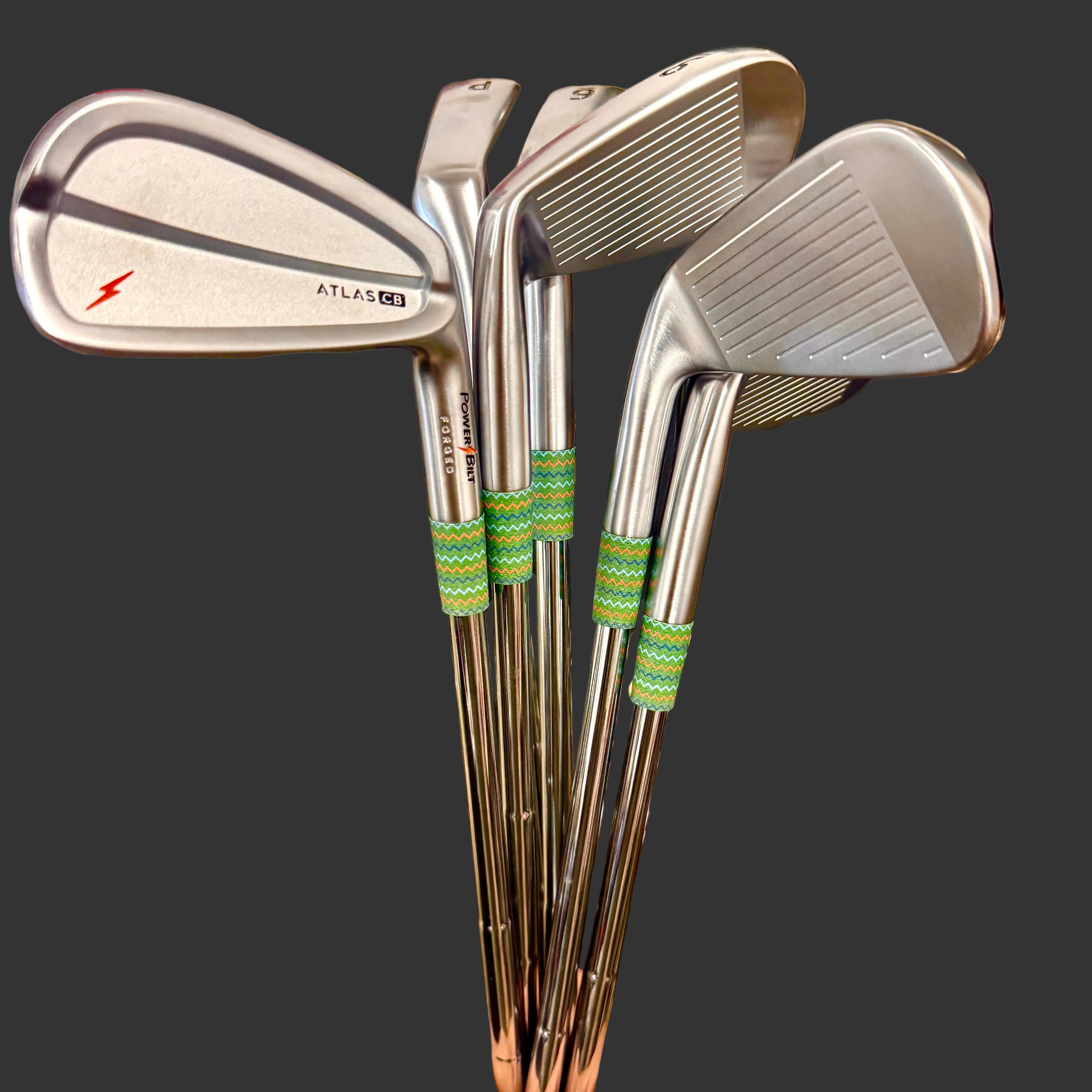 A hand holding a set of four silver golf irons, including an Atlas B55 iron, in front of a black mat with gold script logos.