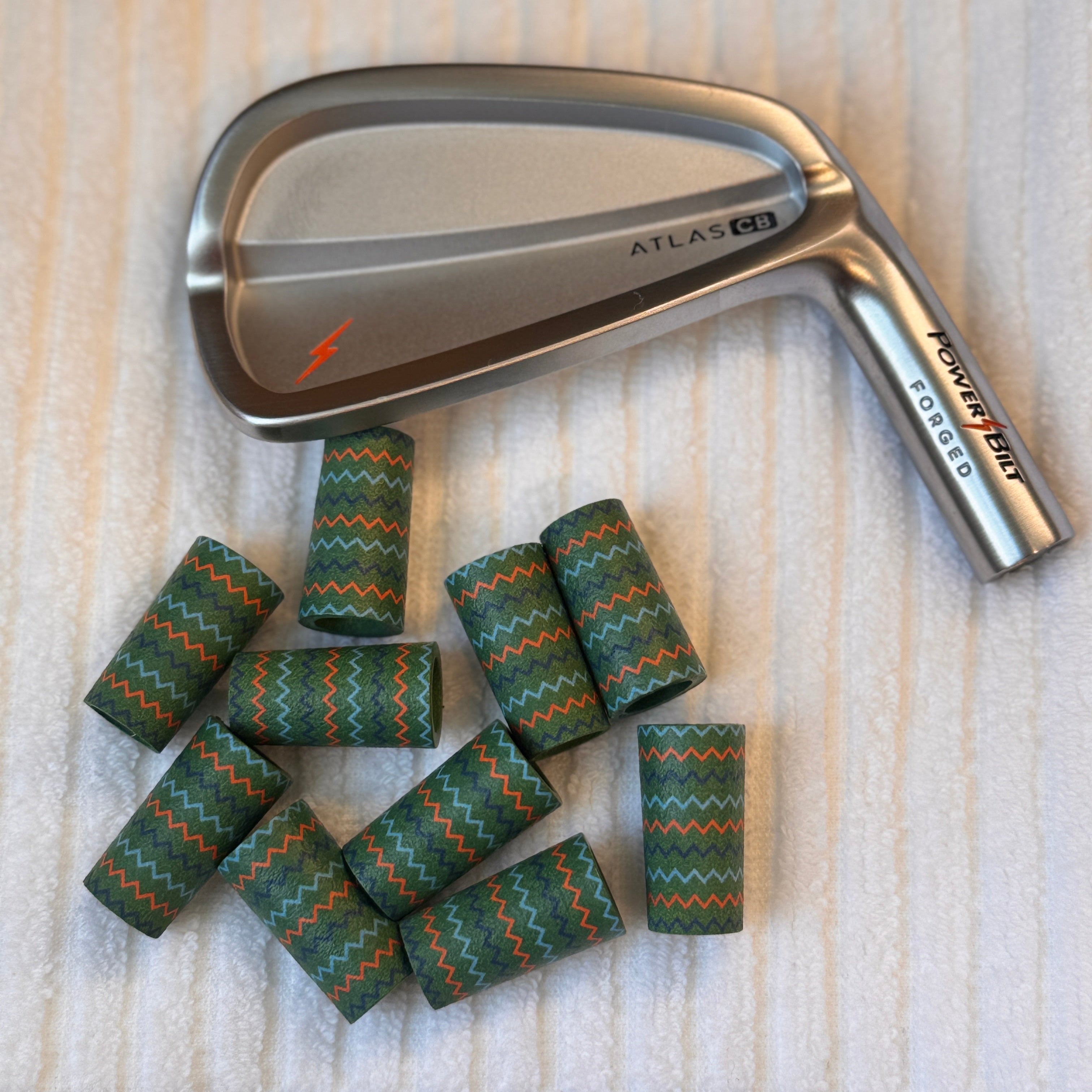 A silver Powerbilt Atlas B1 golf iron club shown on a white textured towel, with a small pile of green and orange chevron-patterned golf ferrules in front.