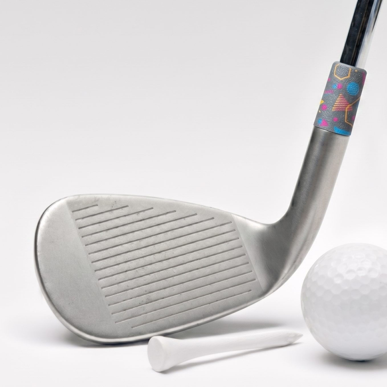 A silver golf club head with a colorful patterned ferrule, shown alongside a white golf ball, tees, and a circular inset close-up of the grip detail on a clean white background.