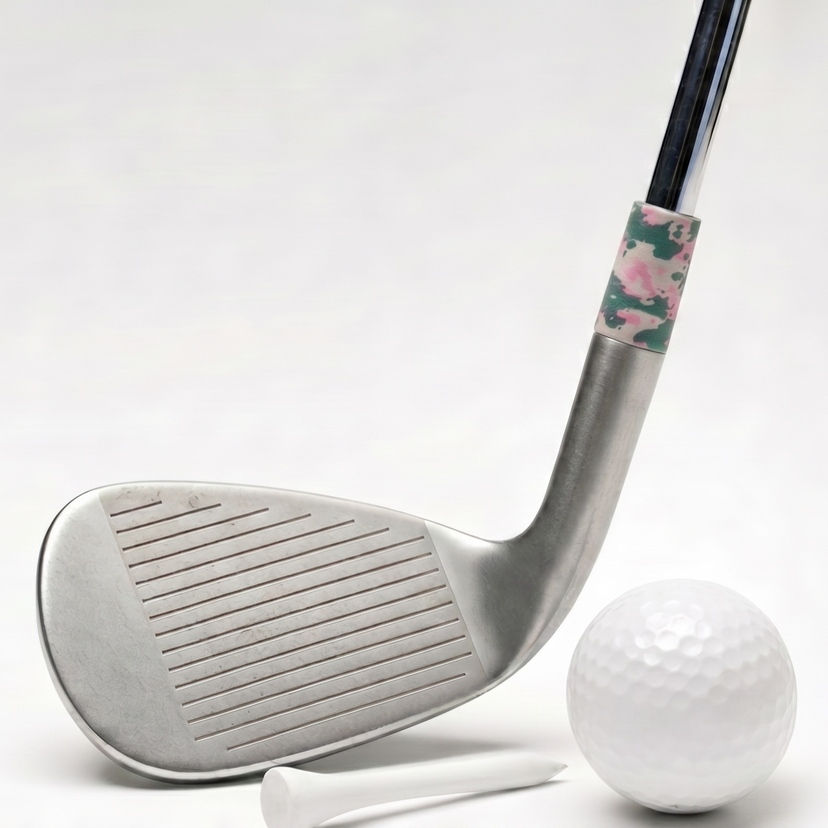 A silver golf club head with a black shaft and a pink and green camouflage ferrule, shown alongside a white golf ball, tees, and a circular inset close-up of the camo ferrule detail.