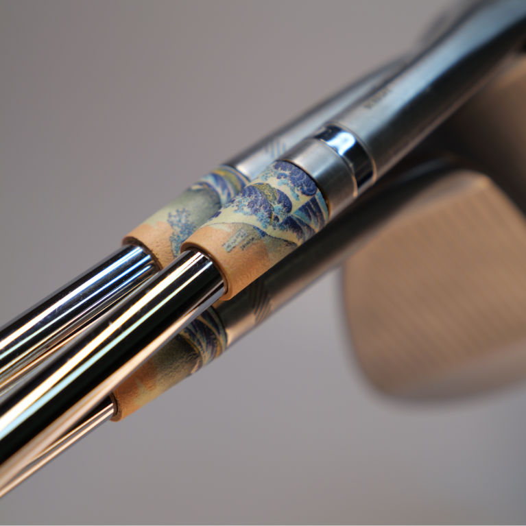 Close-up of a golf club head and shafts featuring a decorative wave-patterned ferrules in blue, beige, and gold tones, with a blurred neutral background.