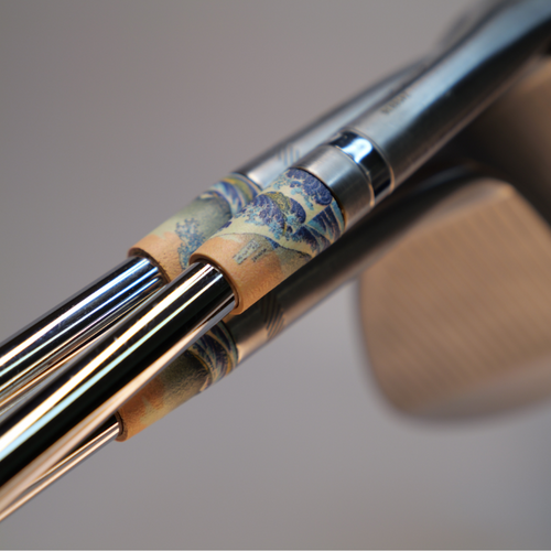 Close-up of a golf club head and shafts featuring a decorative wave-patterned ferrules in blue, beige, and gold tones, with a blurred neutral background.